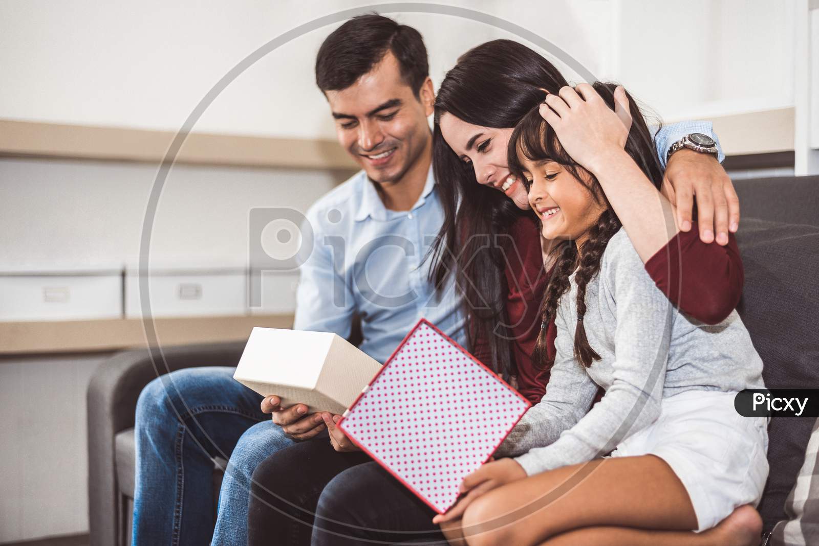 Happy Family Parents And Little Girl Looking Into Gift Box In Christmas And New Year Day On Sofa In Living Room. Xmas Present For Surprised Good Children In Happy Home. People And Lifestyles Concept.