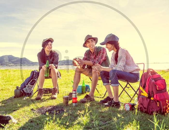 Group Of Campers Discussing About Picnic At Meadow Field With Mountain And Lake Background. People And Lifestyles Concept. Hiking And Travel Theme. Three People With Backpack.