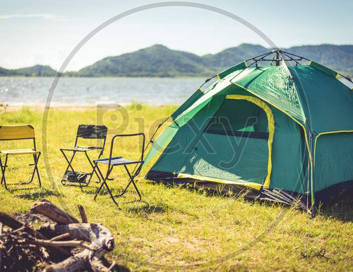 Camping Tent With Extinguished Bonfire In The Green Field Meadow, Lake And Mountain Background. Picnic And Travel Concept. Nature Theme.