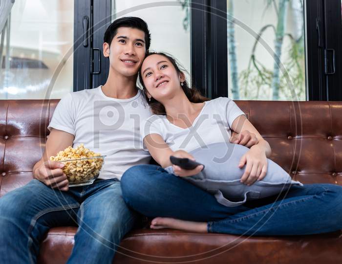 Asian Couples Watching Television And Eating Popcorn Together On Sofa In Their Home. People And Lifestyles Concept. Happy Home And Activity Theme.