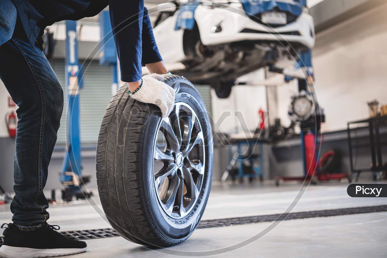 Male Mechanic Hold And Rolling Tire At Repairing Service Garage Background. Technician Man Replacing Winter And Summer Tyre For Safety Road Trip. Transportation And Automotive Maintenance Concept
