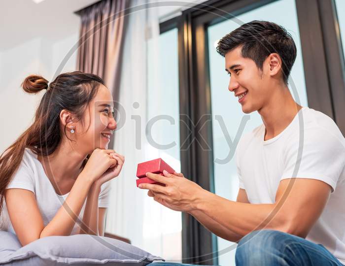 Man Holding Gift Box For Surprise Girlfriend At Their Home. Woman Waiting For Valentines Gift Form Boyfriend. Happy Birthday Party Anniversary Concept. People Couple Lifestyles And Family Life Theme