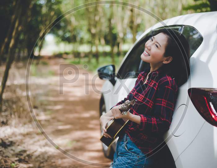 Beauty Asian Woman Smiling And Having Fun At Outdoors Summer With Ukulele Near White Car. Traveling Of Photographer Concept. Hipster Style And Solo Woman Theme. Lifestyle And Happiness Life Theme.