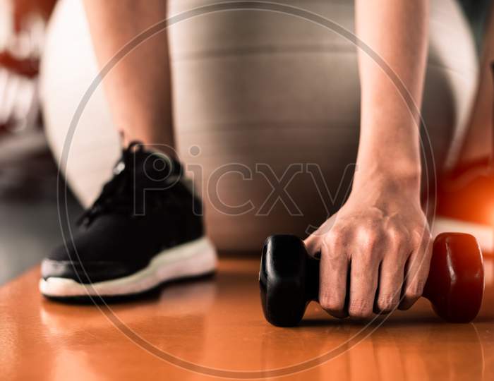 Close Up Of Sport Woman Sitting On Yoga Ball And Grab Dumbbell On Floor By Hand In Fitness Sport Club Center Background. Sport And Recreation Activity Concept. Weight Training And Bodybuilding Concept