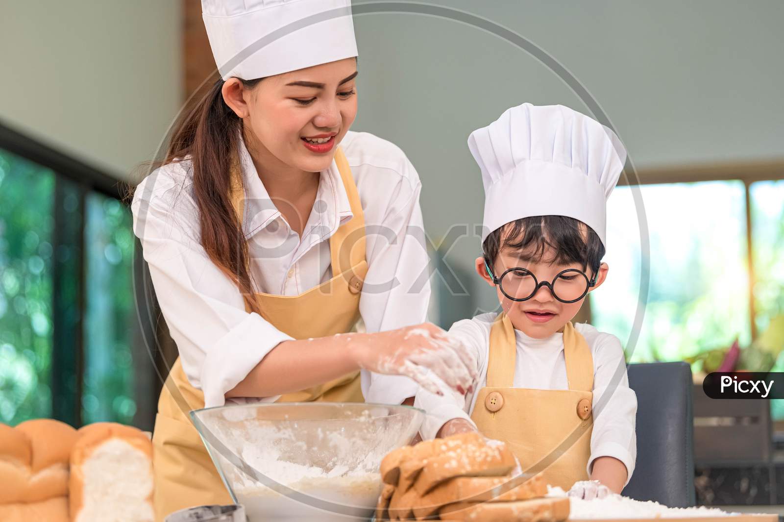 Beautiful Woman And Cute Little Asian Boy With Eyeglasses, Chef Hat And Apron Playing And Baking Bakery In Home Kitchen Funny. Homemade Food And Bread. Education And Learning Concept. Thai Person