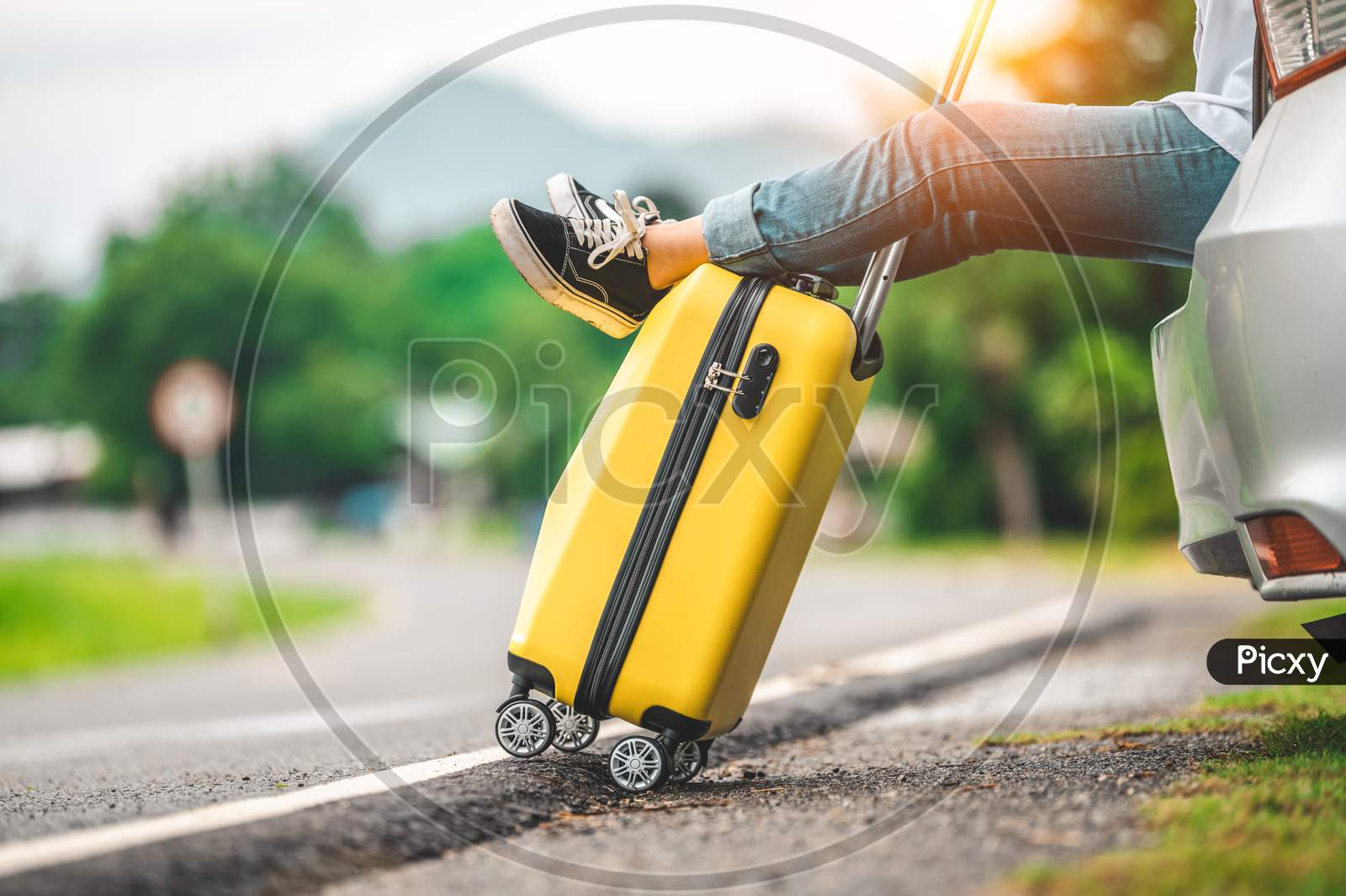 Closeup Of Yellow Luggage And Woman Legs Relaxing On Back Of Car With Road Background. Road Trip And Holiday Vacation Concept. People Lifestyles And Transportation. Girl Spending Weekend In Roadtrip