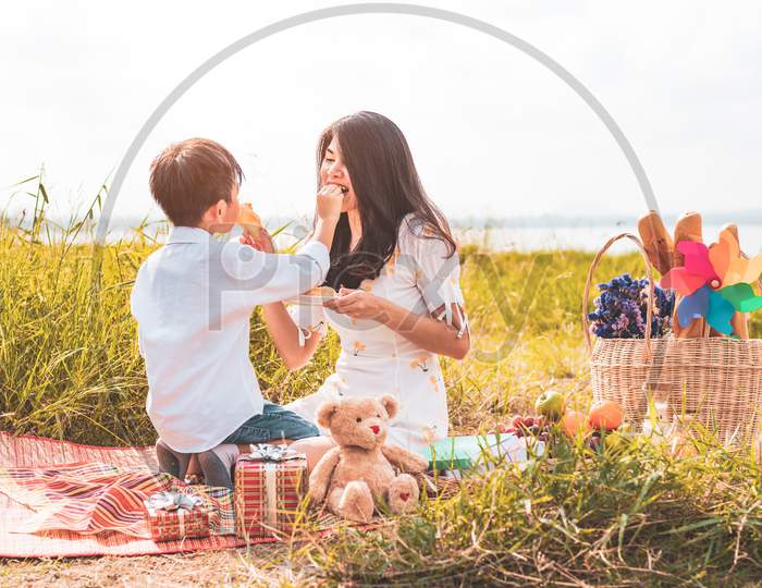 Little Asian Boy His Mom Feeding Snack Each Other In Meadow When Doing Picnic. Mother And Son Playing Together. Celebrating In Mother Day And Appreciating Concept. Summer People And Lifestyle Theme.