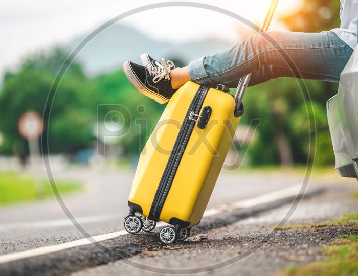 Closeup Of Yellow Luggage And Woman Legs Relaxing On Back Of Car With Road Background. Road Trip And Holiday Vacation Concept. People Lifestyles And Transportation. Girl Spending Weekend In Roadtrip