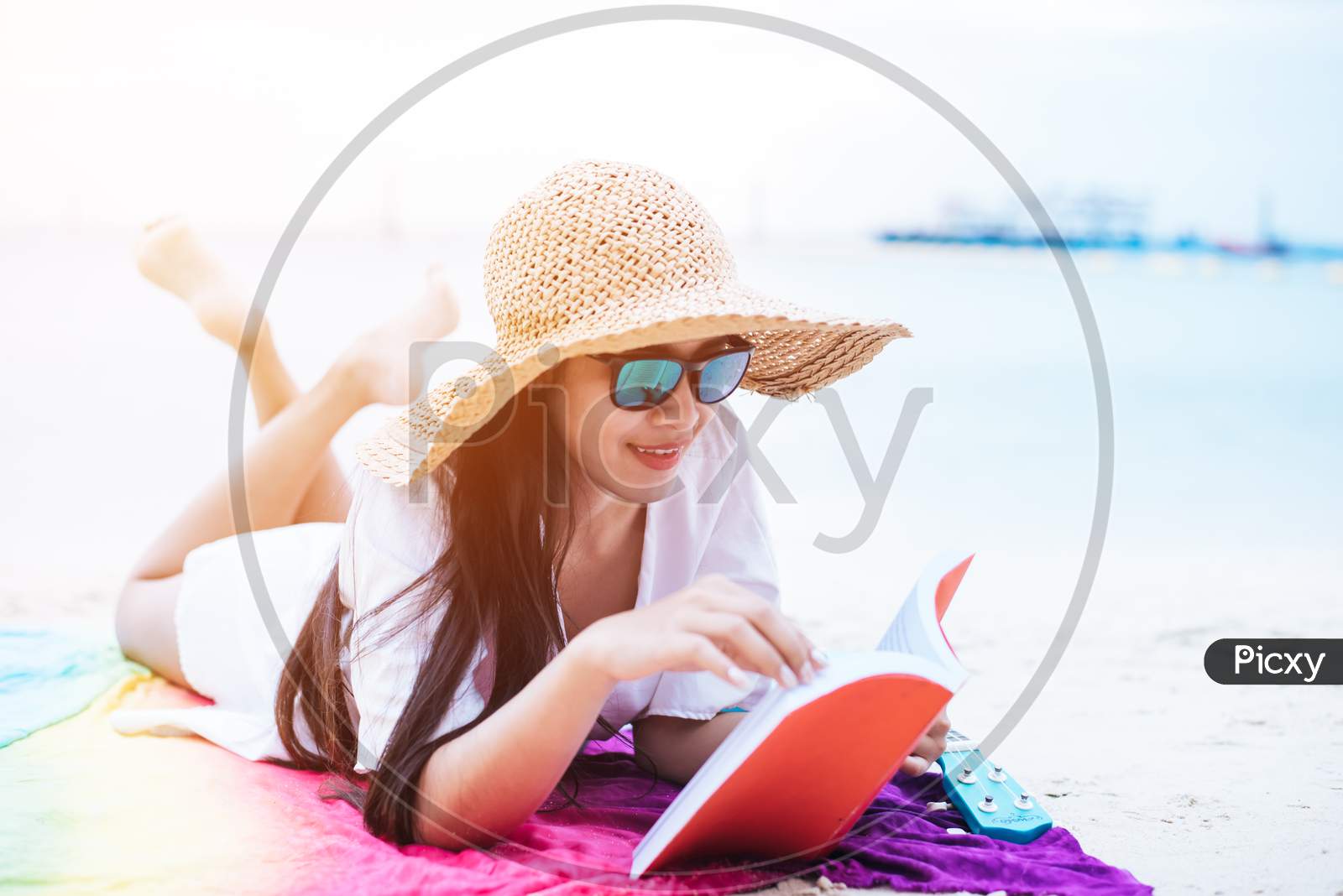 Beauty Asian Woman Have Vacation On Beach. Girl Wearing Wing Hat And Reading Book On Colorful Mat Near Sea. Lifestyle And Happy Life Concept. Travel And Holiday Theme.