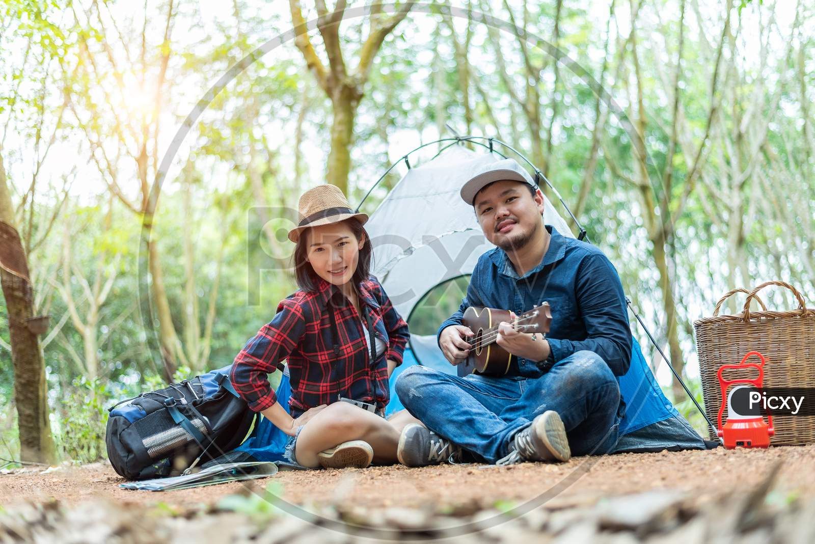 Asian Couple Camping In The Forest. Man Playing Ukulele With Woman In Front Of Camping Tent. Lamp And Basket And Backpack Element. People And Outdoor Lifestyles Concept. Adventure And Travel Theme.