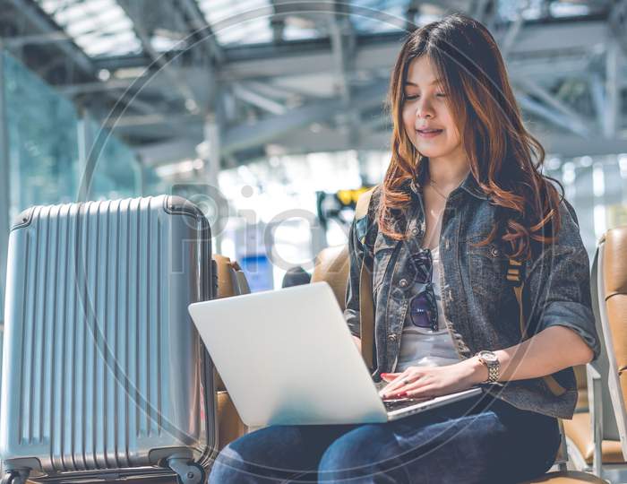 Beauty Asian Woman Using Laptop With Suitcase While Waiting For Take Off Flight. Woman Sitting In Airport Terminal. People And Lifestyle Concept. Technology And Travel Theme. Portrait Theme