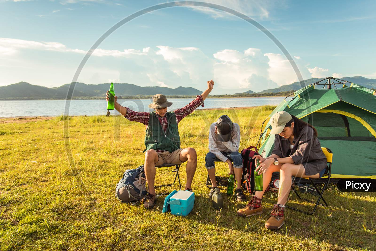 Drunken Tourists Doing Party While Camping And Picnic In Meadow Field. Mountain And Lake Background. People And Lifestyles Concept. Outdoors Activity And Leisure Theme. Backpacker And Hiker Theme