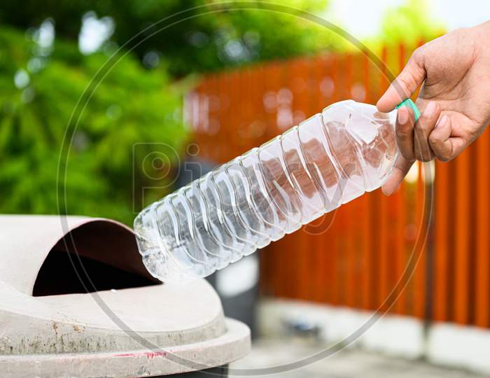 Closeup Of Hand Throwing Away Empty Plastic Bottle Into Trash For Recycling In Front Of House Background. World Environment Day Concept.
