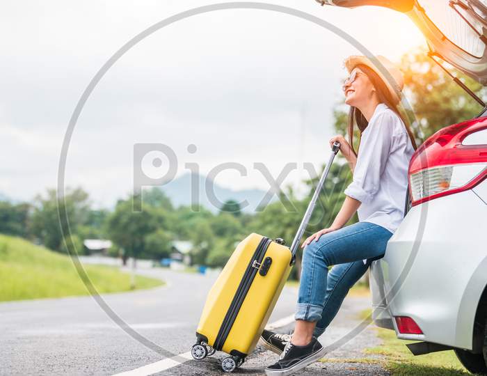 Asian Woman Spending Weekend In Roadtrip With Yellow Luggage. Girl Relaxing On Back Of Car With Road Background. Road Trip And Holiday Vacation Concept. People Lifestyles And Transportation.