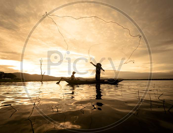 Silhouette Fisherman Fishing By Using Net On The Boat With Sunlight In Thailand, Nature And Culture Concept