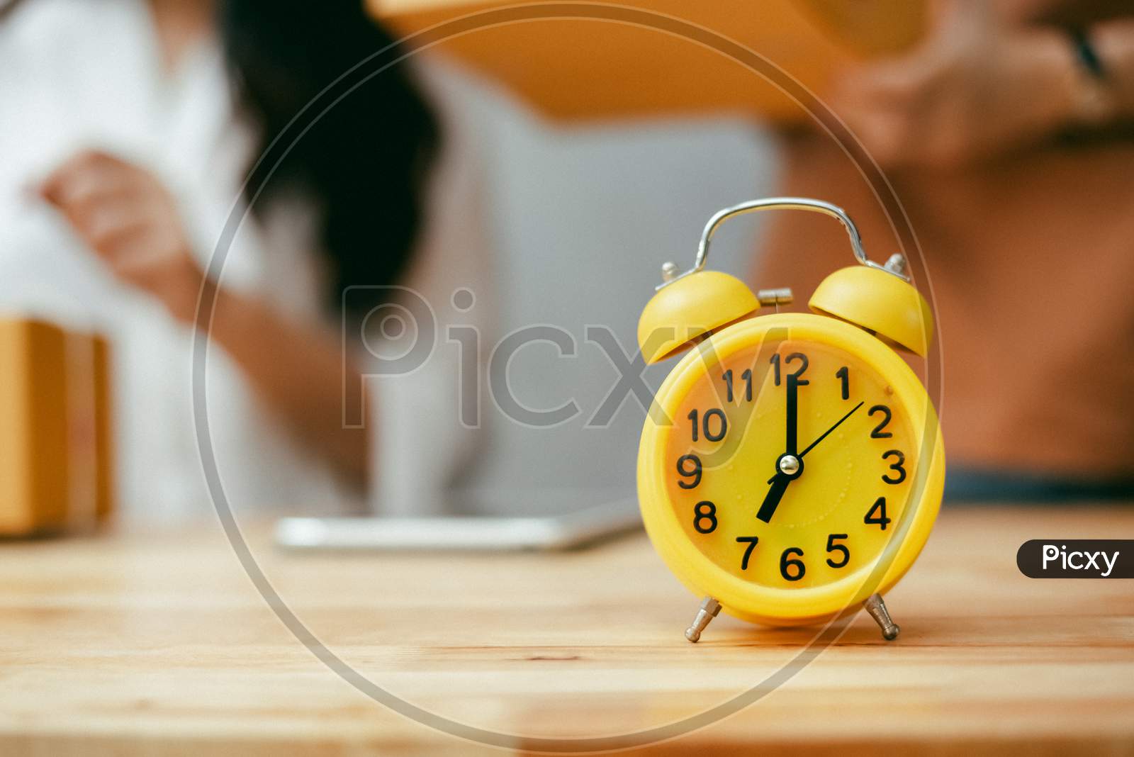Yellow Alarm Clock On Wooden Table In Office. Women Checking Product Stock In Background. Object And Vintage Concept. Home And Office Theme.