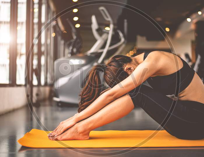 Woman Doing Bending Yoga And Facing Down In Fitness Workouts Training Gym Center. Lifestyle Woman Sitting  With Sport Equipment And Exercise Treadmill Background. Female Yoga Girl In Black Sports Bra