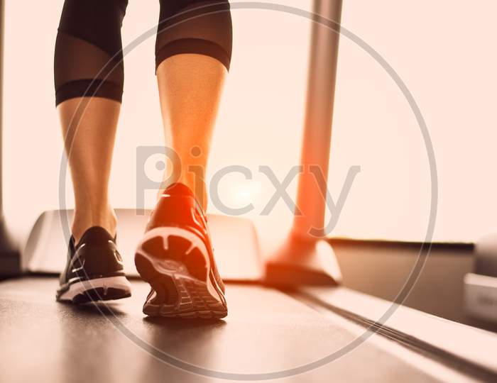 Close Up Of Woman Legs Jogging On Treadmill With Sportwear And Sneakers. Sport And Workout Concept. People And Leisure Concept. Healthcare And Lifestyles Theme. Back View Of Lower Body . Warm Tone