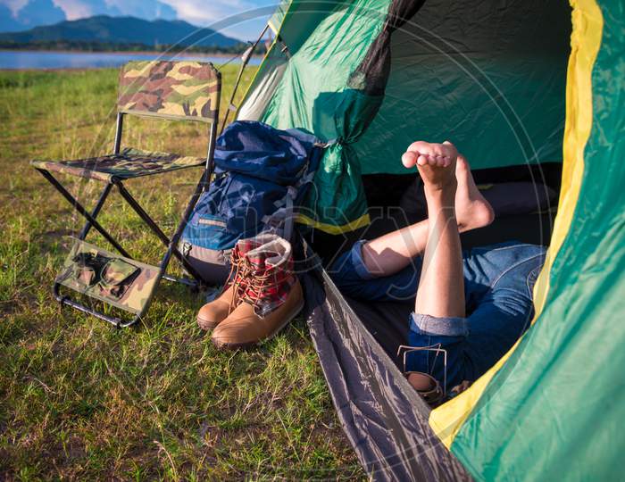 Close Up Of Woman Legs Relaxing In Camping Tent With Mountain Lake And Meadow And Grass Field Background. Lifestyles And People Concept. Camping And Picnec Theme. Green Natural And Summer Travel Theme