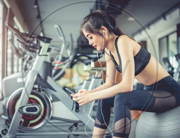 Asian Woman Sitting On Fitness Ball And Listening Music In Fitness Training Gym With Smart Phone. Relax And Sport Workout Training Concept. Technology And Entertainment Theme
