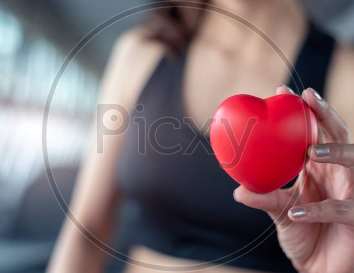 Close Up Of Red Massage Ball Like Heart Shape In Fitness Woman Hand At Sport Gym Training Center. Medical And Healthcare Of Heart Attack And Serious Illness Concept. Female Sporty Girl Wear Sport Bra.
