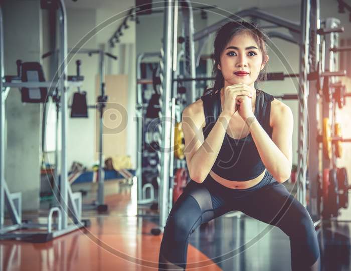 Young Asian Woman Doing Squat Workout For Fat Burning And Diet In Fitness Sports Gym With Sports Equipment In Background. Beauty And Body Build Up Concept. Sports Club And Aerobic Theme.