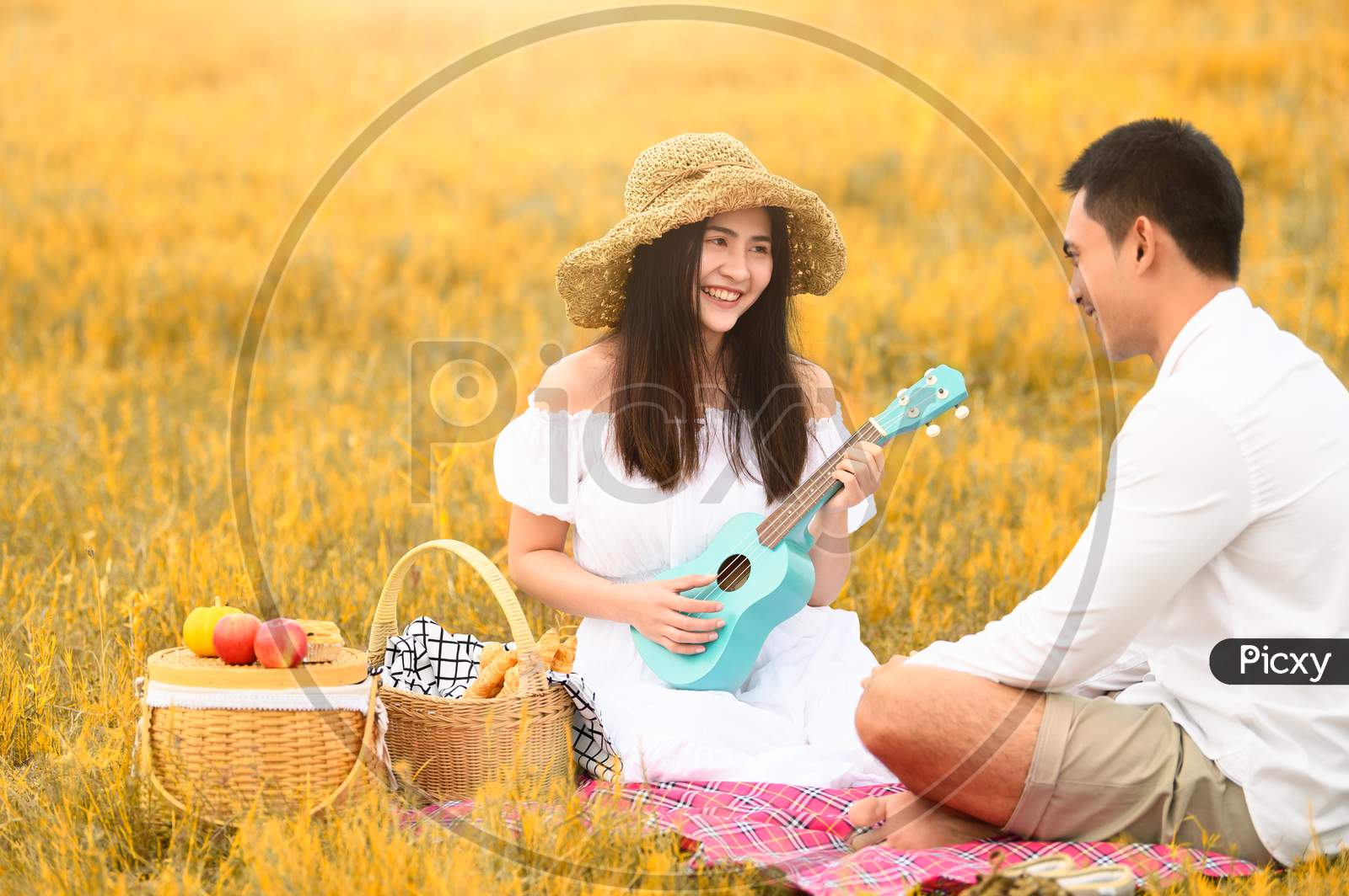Two Asian Young Couples In Autumn Meadow Field Doing Picnic In Honeymoon Trip In White Clothes, Ukulele Guitar And Fruits Basket. People Lifestyle And Wedding Concept. Nature And Travel Day Concept.
