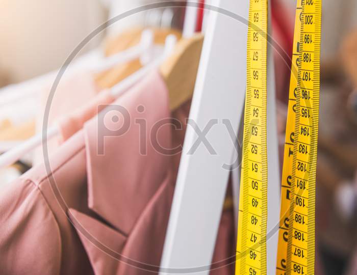 Closeup Of Measuring Tape And Line Of Fashion Pastel Pink Colored Woman Shirts With Wooden Hangers In Clothing Shop.