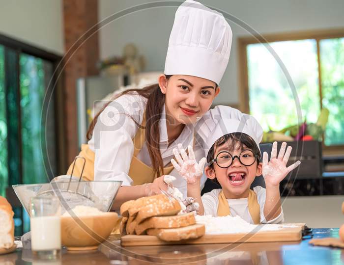 Beautiful Woman And Cute Little Asian Boy With Eyeglasses, Chef Hat And Apron Playing And Baking Bakery In Home Kitchen Funny Concept. Homemade Food And Bread. Funny Face Emotion Looking Camera