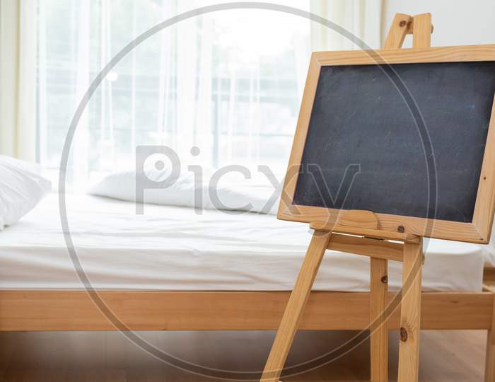 Black Chalkboard On Easel Stand In Bedroom With White Soft Bed Background. Interior And Decoration Concept.