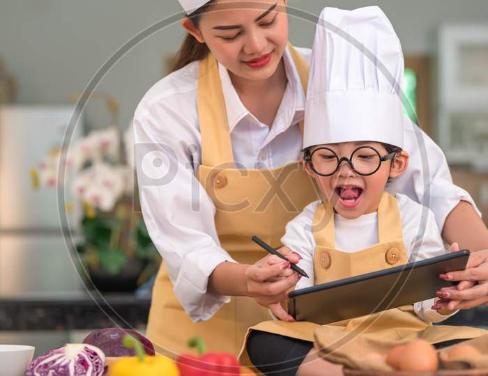 Beautiful Asian Woman And Cute Little Boy Prepare Online Shopping And Listing Ingredient For Cooking In Kitchen At Home With Tablet. People Lifestyles And Family. Homemade Food And Ingredient Concept.