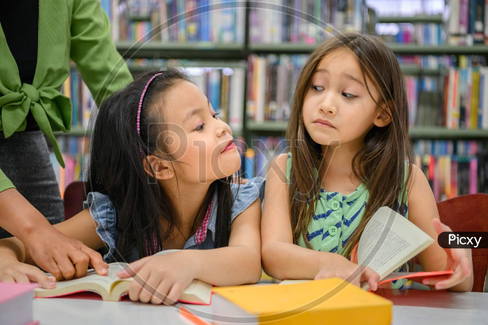Two Cute Girls Are Jealous Of Each Other While Reading Books In Library While Teacher Teaching. People Lifestyles And Education. Young Friendship And Kids Relationship In School Concept. Daycare Theme