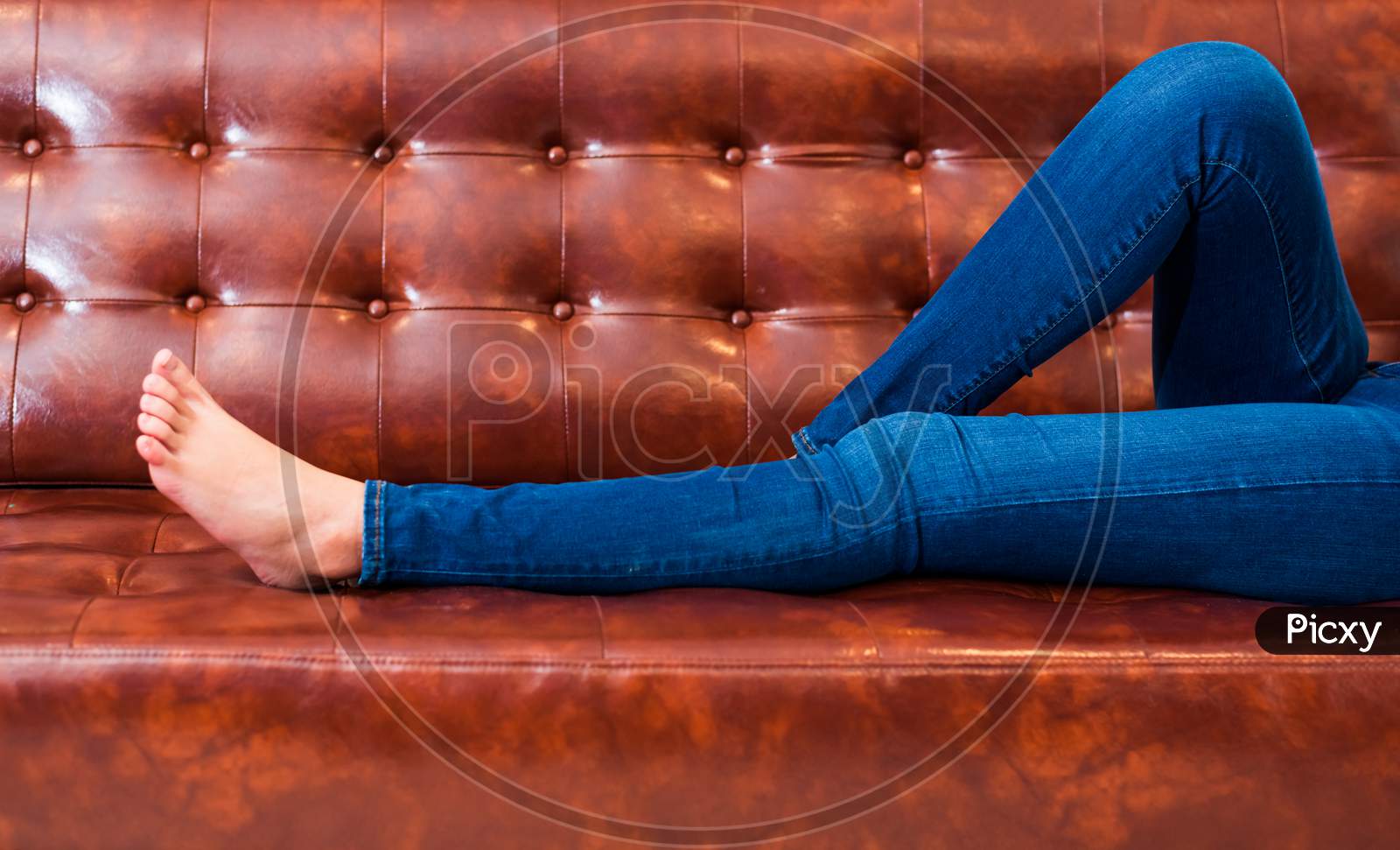 Woman Legs With Wearing Jeans While Relaxing At Movie Theater Or Home On Brown Leather Sofa, Relax And Holiday Concept, Bed Time Sleeping Concept. People Lifestyle And Holiday. Closeup Girl Foot