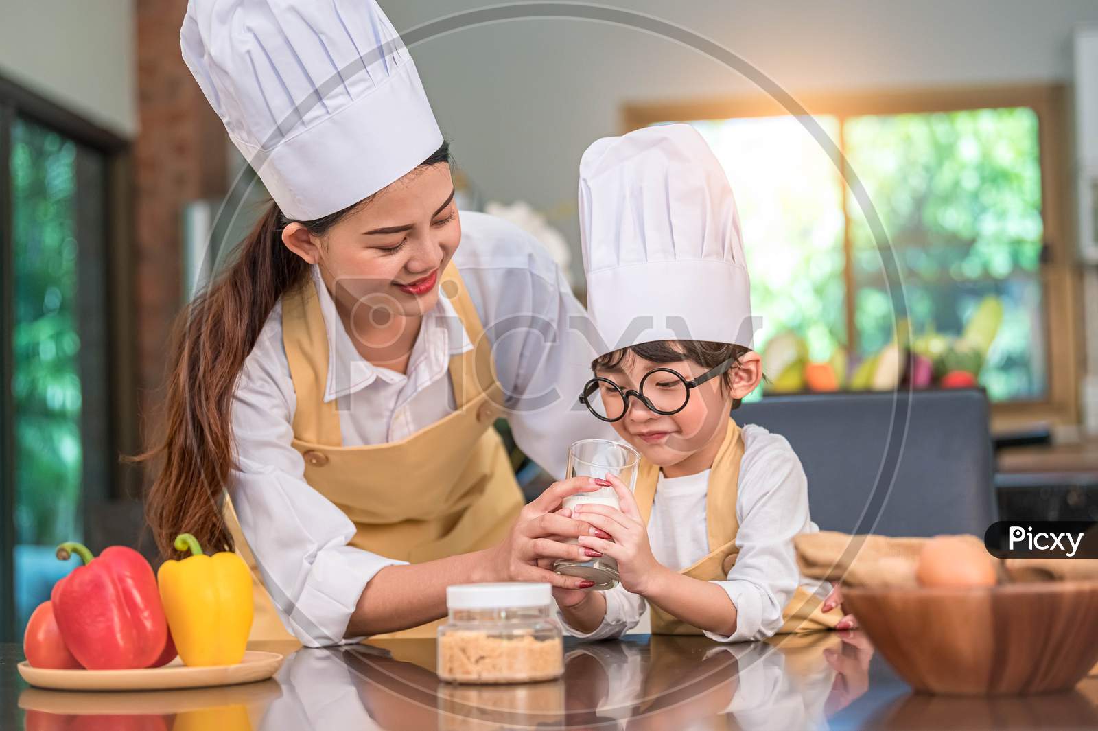 Beautiful Asian Woman Teaching Cute Little Boy With Eyeglasses To Drink Milk In Kitchen At Home Together. Lifestyles And Family. Son Dislike Milk And Doing Funny Face With Milk Glass. Food And Drink