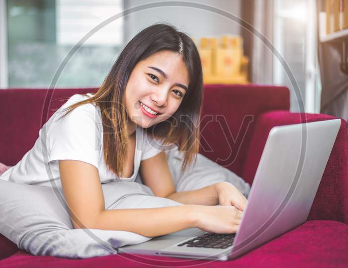 Young Woman Surfing The Internet By Laptop On Red Sofa In Cheerful Gesture Mood Emotion. Selling And Online Shopping Concept. Merchant And Marketing. Happiness Of New Business Trader  And Entrepreneur