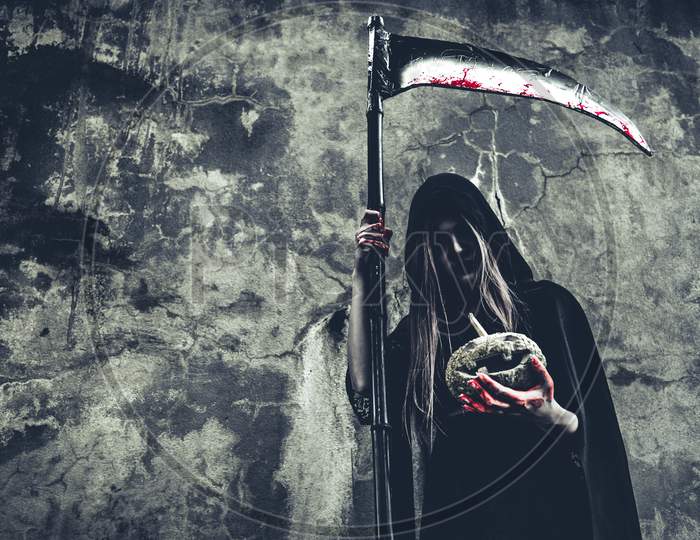 Demon Witch Holding Pumpkin In Hand With Bloody Reaper On Grunge Wall Background. Halloween And Religious Concept. Demon Angel And Satan Theme.