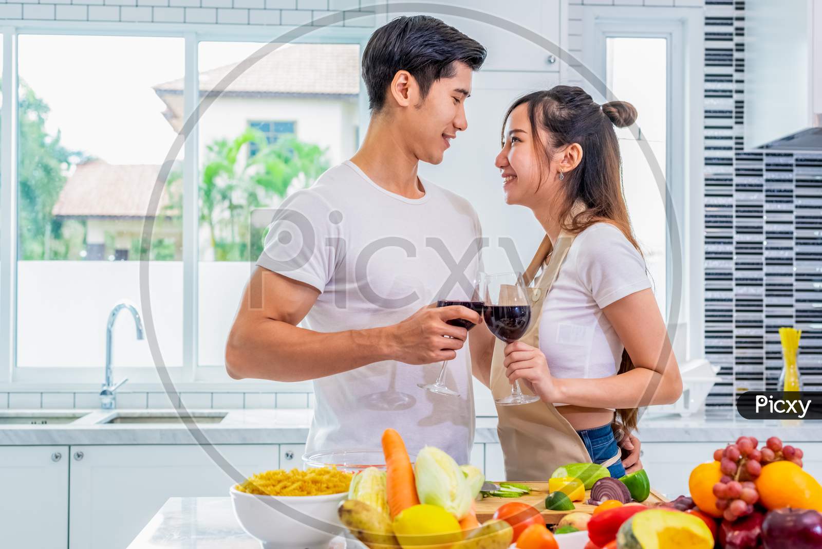 Happy Asian Young Married Couple Drinking Wine And Looking Each Other In Home Kitchen. Boyfriend And Girlfriend Cooking Together. People Lifestyle And Romantic Relationship Concept. Valentines Day