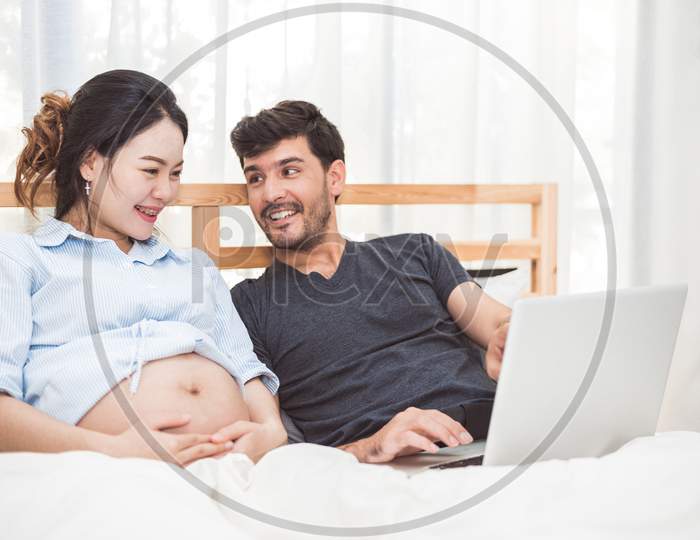 Happy Man And His Pregnant Wife Using Laptop To Searching Newborn Baby Items For Preparing Parenthood. Couple Lifestyle Family And Technology Concept. Women Health And Medical Theme.