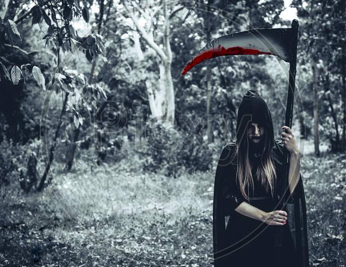 Female Demon Witch With Bloody Reaper Standing In Front Of Mystery Forest Background. Halloween And Religious Concept. Demon Angel And Satan Theme.