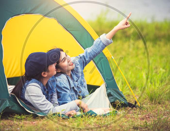 Two Close Friend Asian Friendship Relax In Camping Tent In Green Meadow On Lake Side View Background. Girl Pointing Finger To Sky. People Lifestyle Travel On Vacation Concept. Summer Picnic Activity