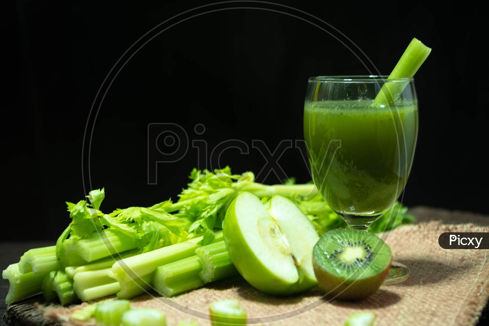 Blended Mixed Juice Celery And Kiwi And Green Apple In Welcome Drink Glass And Bunch Of Fresh Celery Stalk On Wooden Table With Leaves On Black Background. Food And Ingredients  Of Healthy Vegetable.