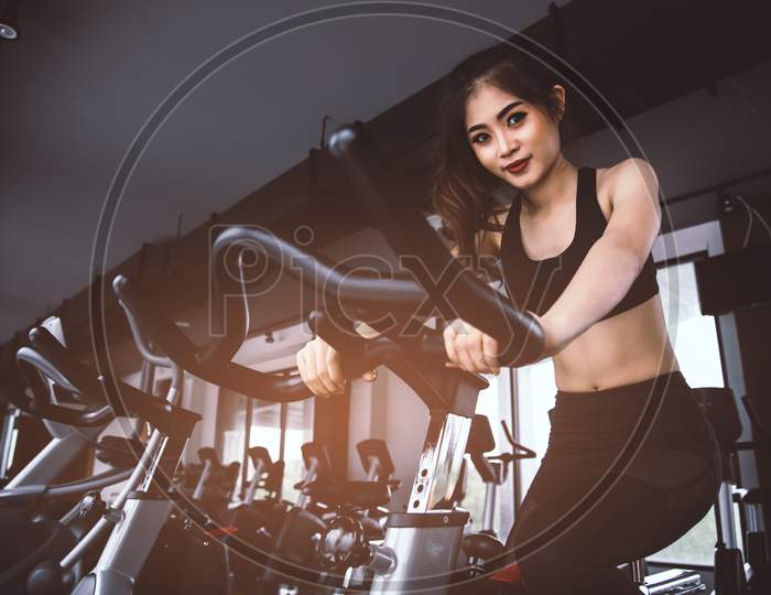 Asian Fitness Woman On Bicycle Doing Spinning At Gym. Beautiful Young Female Work Out On Gym Bike. Medical And Healthy Concept. Workout And Cardio Theme.