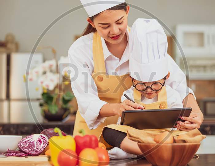 Beautiful Asian Woman And Cute Little Boy Prepare Online Shopping And Listing Ingredient For Cooking In Kitchen At Home With Tablet. People Lifestyles And Family. Homemade Food And Ingredient Concept.