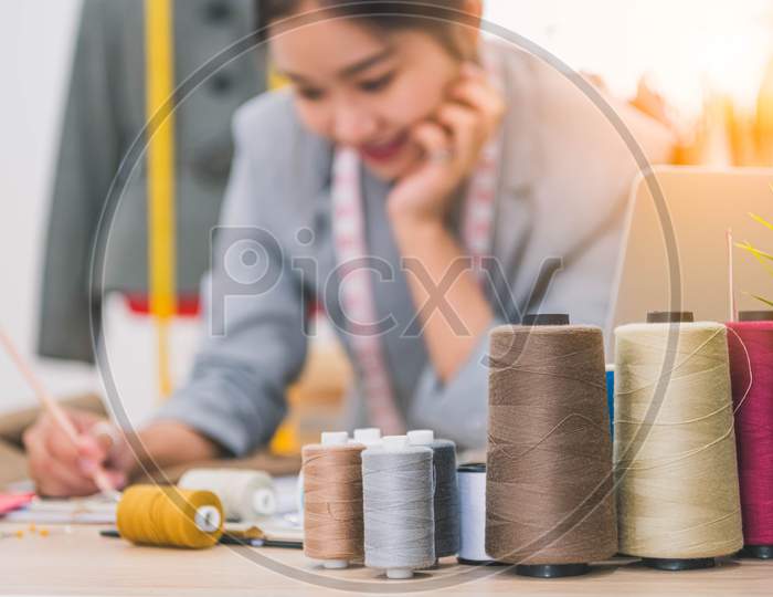 Closeup Of Color Thread With Blurry Happy Female Tailor And Sewing Clothing Stylist Designer Background. Woman Creating New Collection In Workshop Office. People Lifestyle And Freelance Occupation Job