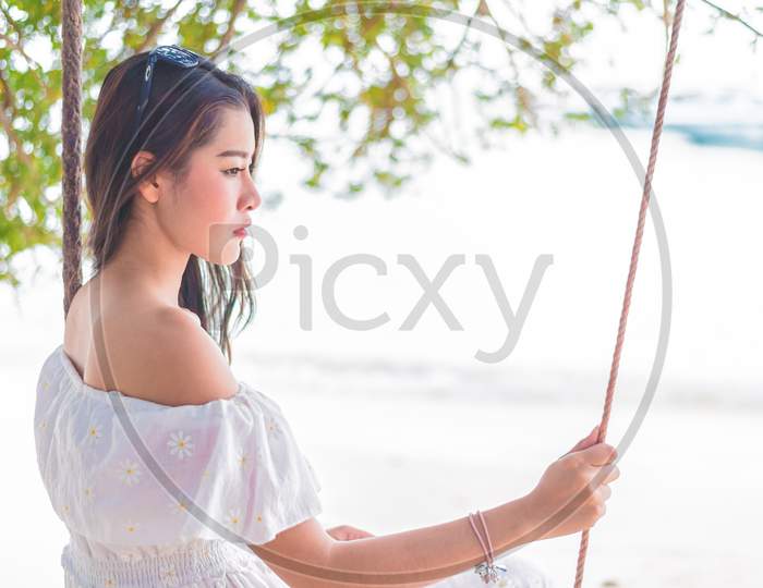 Asian Woman On White Dress Sitting On Swing At Beach. People And Nature Concept. Sad Love And Missing Someone Concept. Lonely And Heart Broken Theme.