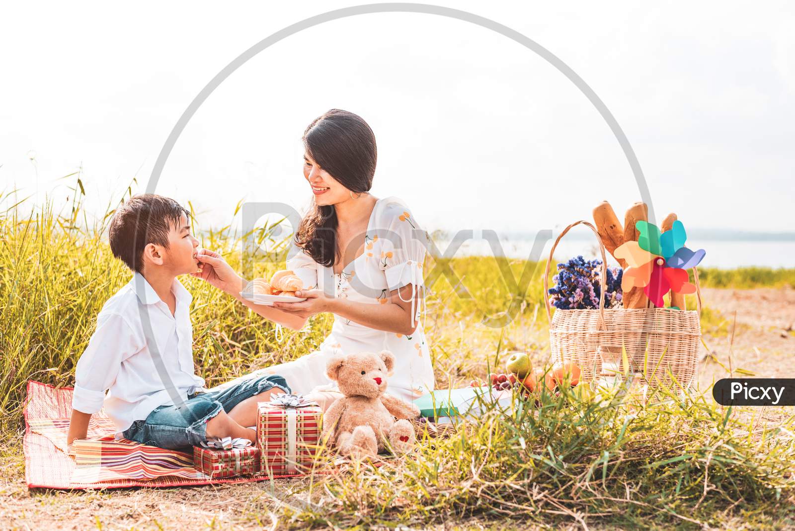 Beautiful Asian Mom Feeding Snack To Her Son In Meadow When Doing Picnic. Mother And Son Playing Together. Celebrating In Mother Day And Appreciating Concept. Summer People And Lifestyle Theme.