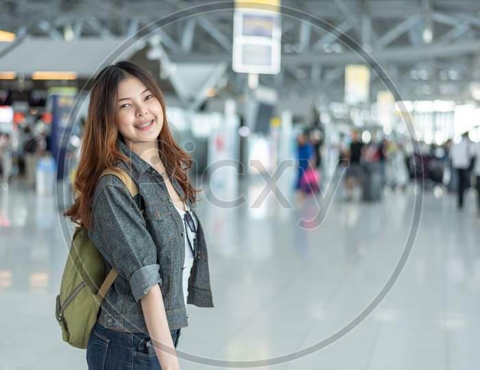 Happy Beauty Asian Woman Traveling And Holding Suitcase In Airport Terminal With Crowd People Passengers Background. People And Lifestyle Concept. Journey Around The World Theme. Cheerful Tourist Girl