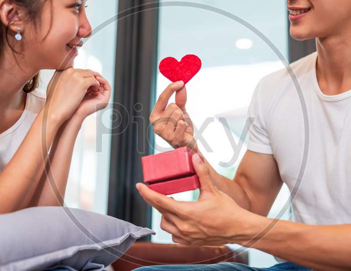 Man Holding Gift Box For Surprise Girlfriend At Their Home. Woman Waiting For Valentines Gift Form Boyfriend. Happy Birthday Party Anniversary Concept. People Couple Lifestyles And Family Life Theme