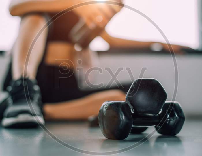 Close Up Of Dumbbell With Exercise Woman Lifestyle Workout In Gym Fitness Breaking Relax After Sport Training With Protein Shake Bottle Background. Healthy Lifestyle Bodybuilding Athlete Muscles