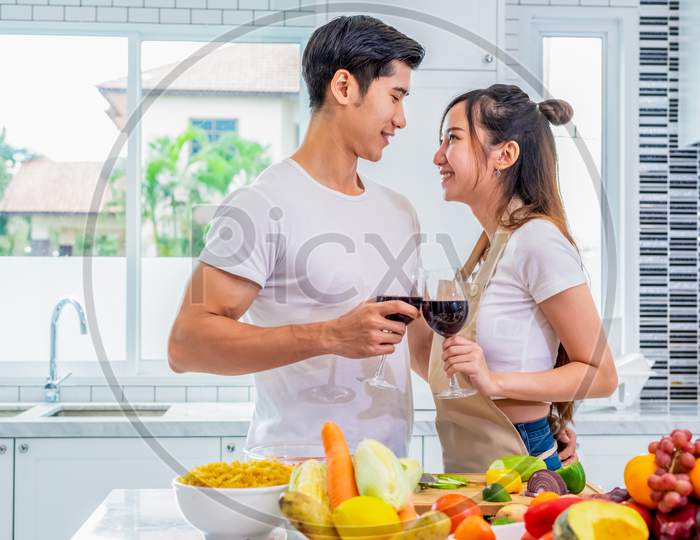 Happy Asian Young Married Couple Drinking Wine And Looking Each Other In Home Kitchen. Boyfriend And Girlfriend Cooking Together. People Lifestyle And Romantic Relationship Concept. Valentines Day